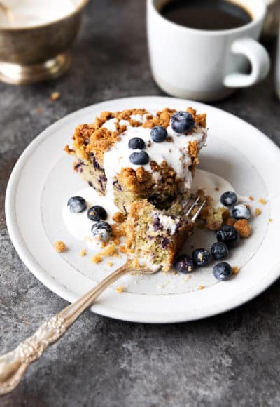 Sour cream blueberry coffee cake covered with light cream on white plate, coffee in background.