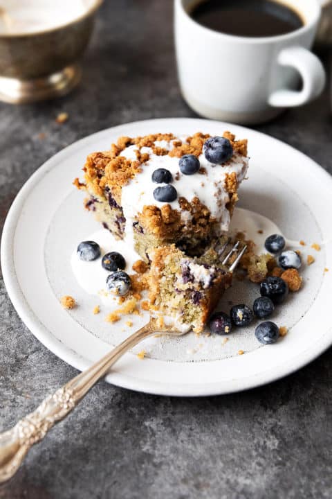 Sour cream blueberry coffee cake covered with light cream on white plate, coffee in background.