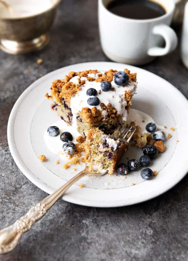 sour cream blueberry coffee cake on white plate, coffee in background. Fork on plate, coffee cake covered with thinned sour cream and blueberries