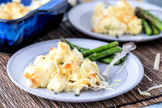 Cauliflower gratin on a white plate with asparagus. More gratin on plate behind and in blue baking dish.