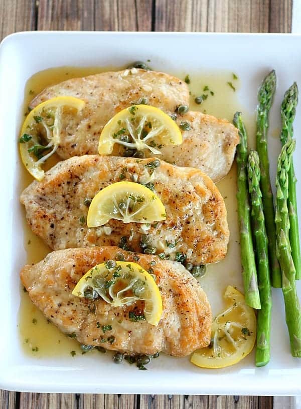Chicken Piccata topped with lemon slices, asparagus on the side on white plate
