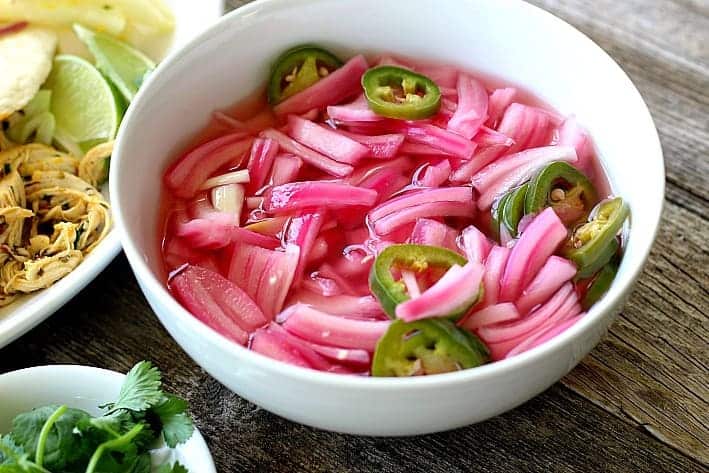 red onions, pickled with jalapenos in a white bowl next to a plate of chicken tacos