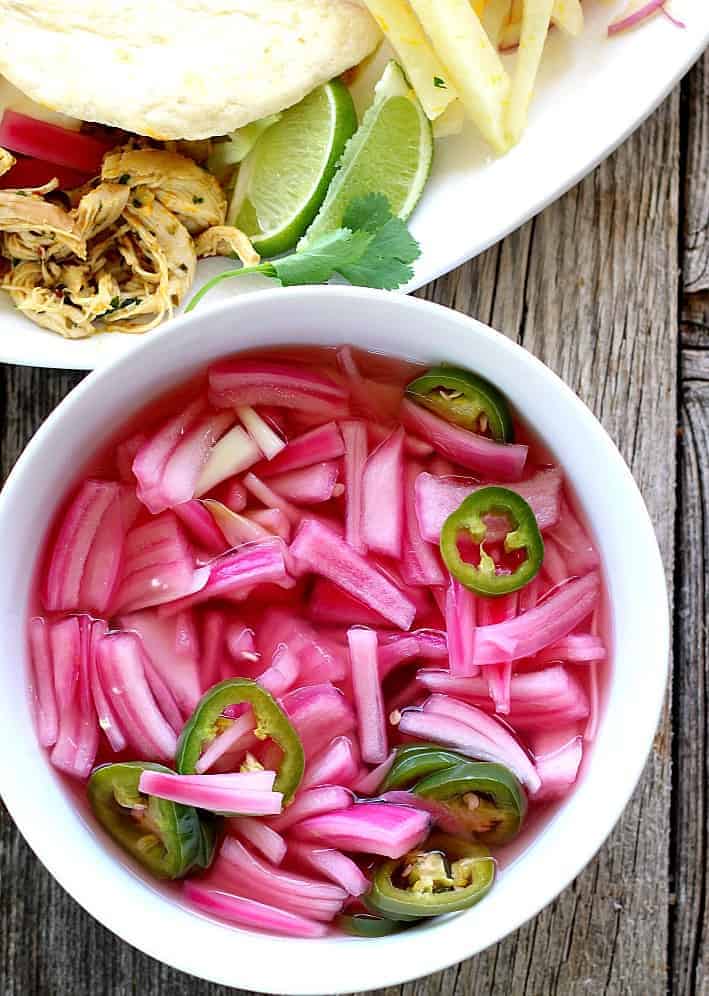 pickled red onions with jalapenos in a white bowl next to a plate of chicken tacos with limes