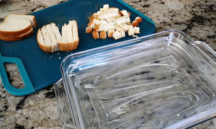Buttered 9x13 pan with sliced, cubed, and whole pieces of bread on a blue cutting board