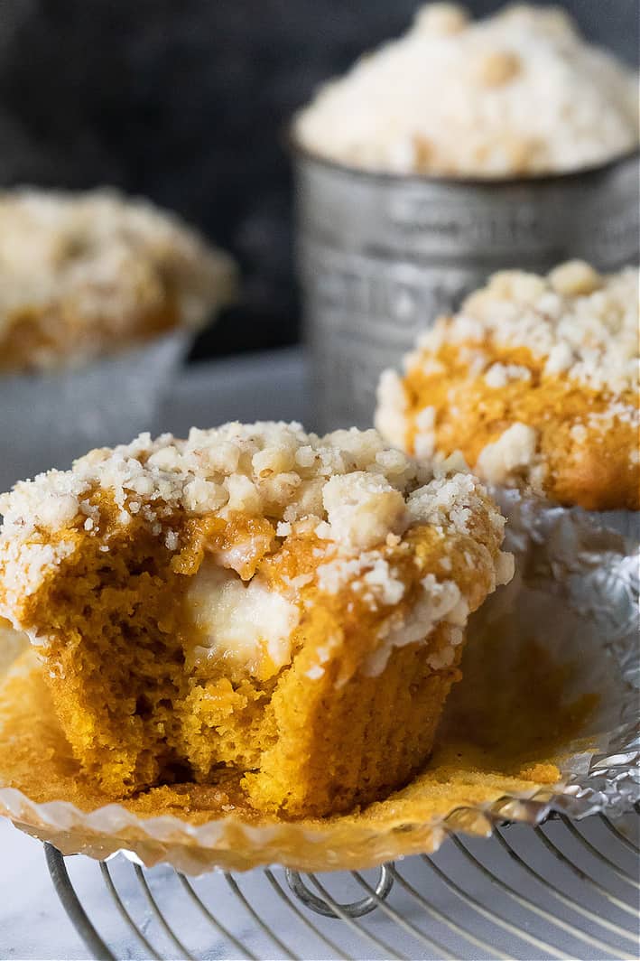 close up of pumpkin muffin with cream cheese with a bite taken out of one. Muffins and a baking tin in the background. Streusel on top