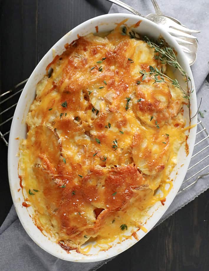 two potato gratin with thyme and shallots in an oblong baking dish with utensils on side. cheese is melted on top.