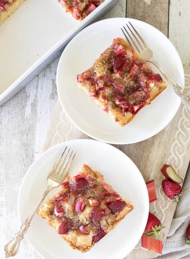 Two slices of Strawberry Rhubarb Kuchen on white plates with forks. Cut strawberries and rhubarb on side board