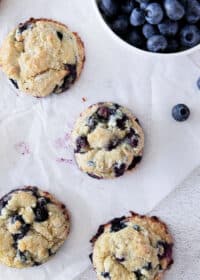 Blueberry muffin cookies on white parchment with blueberries in a bowl above.