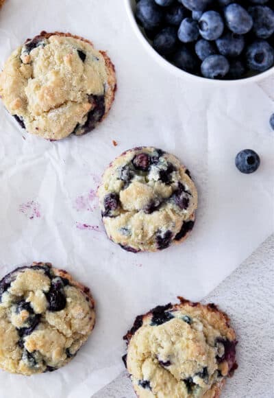 Blueberry muffin cookies on white parchment with blueberries in a bowl above.