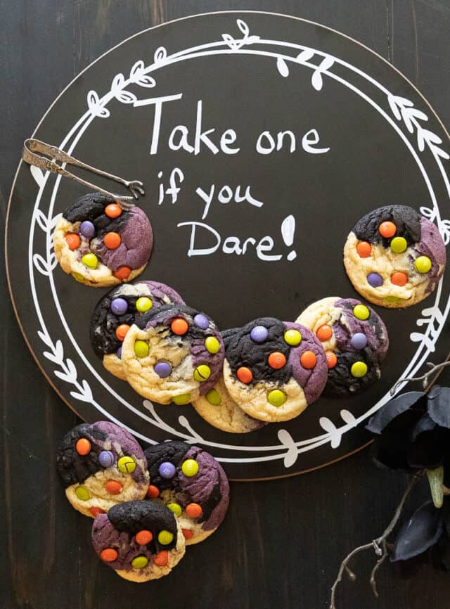 Halloween neapolitan cookies layered on blackboard that says "Take one if you dare". Cookies are purple black and natural with M&M's on them.