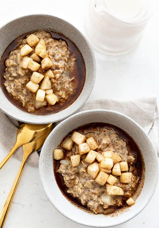two bowls of oatmeal with pears and syrup glaze on top. Two gold spoons and milk on side.