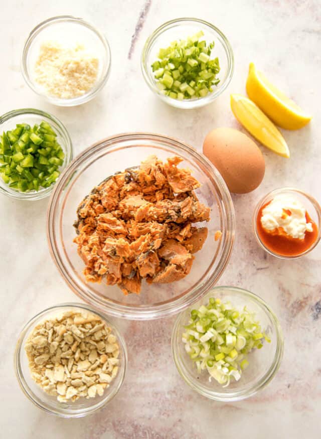 Salmon patty ingredients. Salmon in large bowl in center. Top left and clockwise- Parmesan, celery, lemons and egg, mayonnaise and hot sauce, scallions, crushed saltines, green bell pepper.