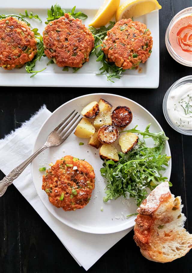 Three salmon patties on rectangle plate at top, pink and dill sauce in small bowls. One salmon patty with potatoes and salad on plate. Bread below plate.