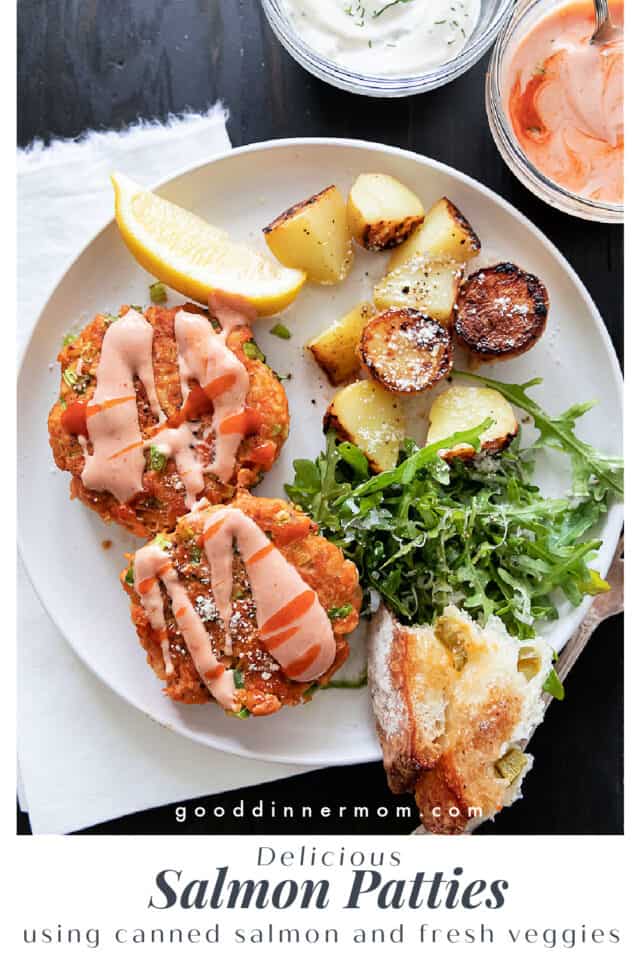 Two salmon patties with pink sauce on white plate with potatoes, salad, bread and lemon. Sauces in small bowls above.