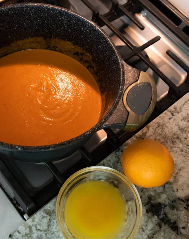 orange tomato bisque in pan with orange juice and orange on counter