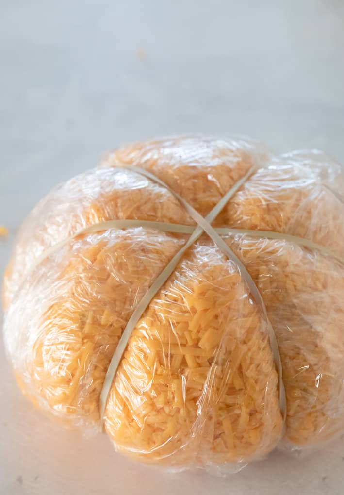 cheese ball wrapped in plastic with three rubber bands wrapped around it to form pumpkin shape