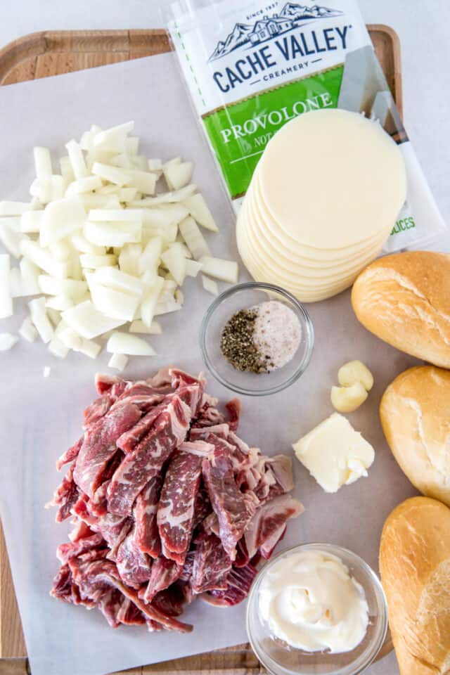 ingredients for philly cheese steaks. Clockwise from top left, chopped onions Cache Valley Provolone, hoagie rolls, mayonnaise, sliced ribeye steak, salt and pepper, garlic and butter.