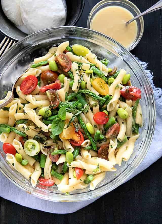 Burrata salad ingredients in a bowl, pasta, asparagus, edamame, tomatoes, with burrata and dressing on side.