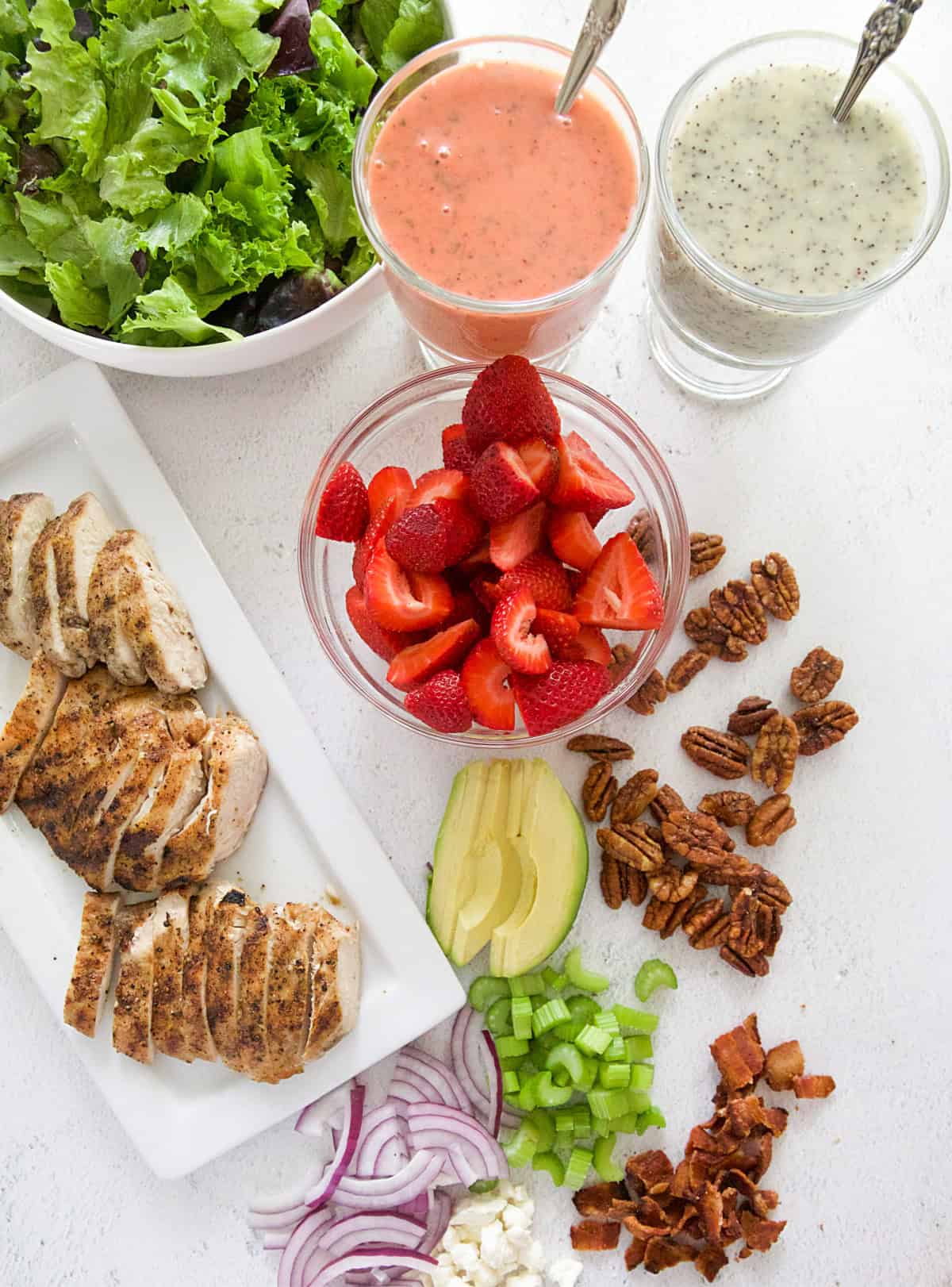 Ingredients for Strawberry Chicken Salad including lettuce, strawberry vinaigrette, poppy seed dressing, strawberries, pecans, avocado, celery, bacon, red onion, feta, and grilled chicken.