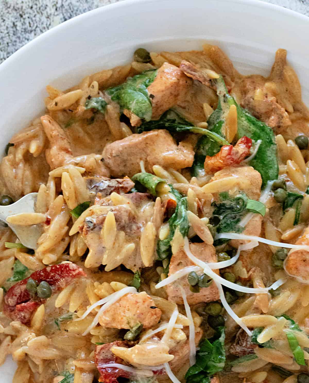 Closeup of One Pot Creamy Chicken Orzo with spinach, sun dried tomatoes, capers, in white bowl,