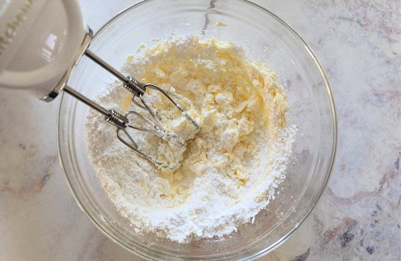 Cream cheese frosting ingredients in bowl with hand held mixer about to fully incorporate.
