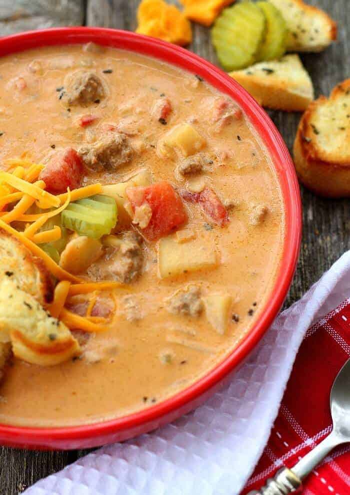 cheeseburger soup in red bowl with croutons, pickle, and cheese on top