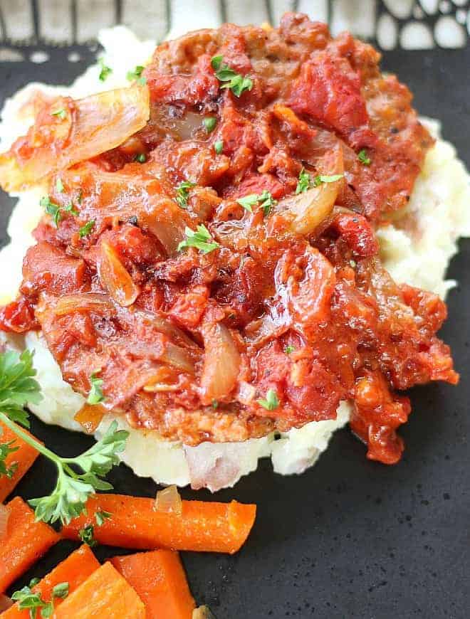 Oven Roasted Swiss Steak on mashed potatoes served on a black plate with cooked carrots on the side