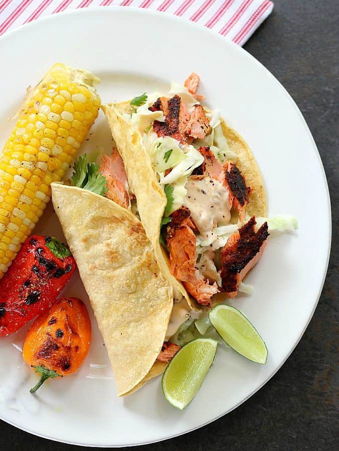 Grilled fish tacos on a white plate served with corn, peppers and lime wedges on the side