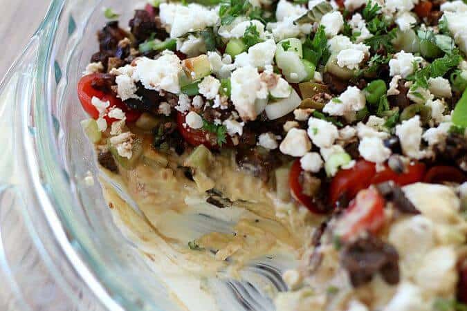 Ultimate Greek 7 Layer Dip shown with portion scooped out. Feta and green onions on top. Kalamata olives, tomatoes and hummus.