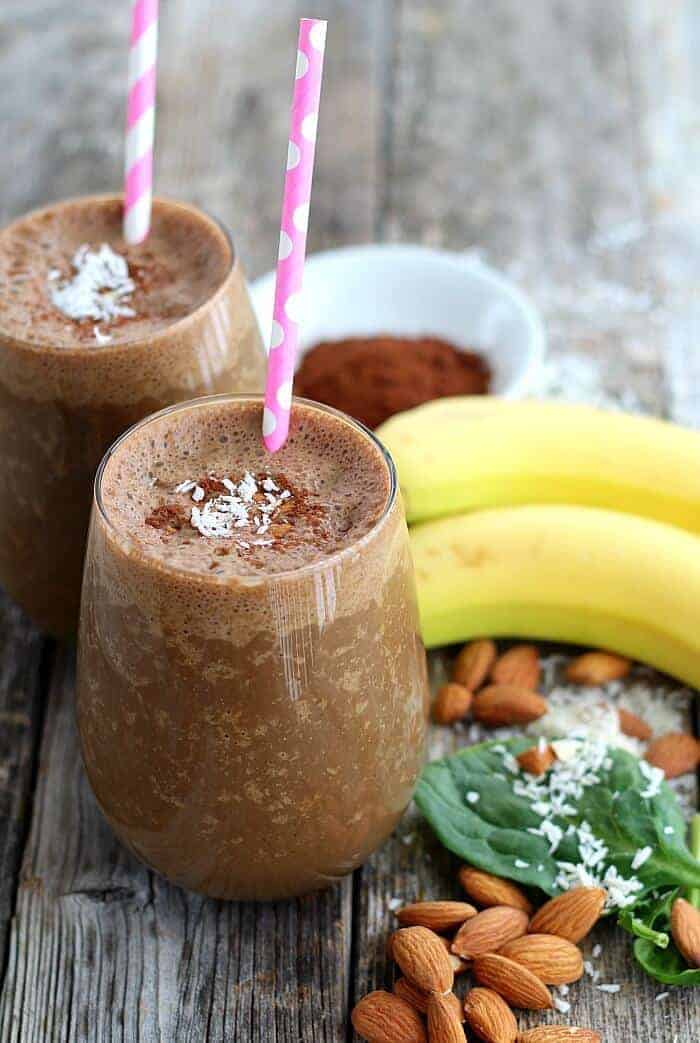 Two glasses of almond joy smoothies with bananas, cocoa powder, almonds, and coconut on the side.
