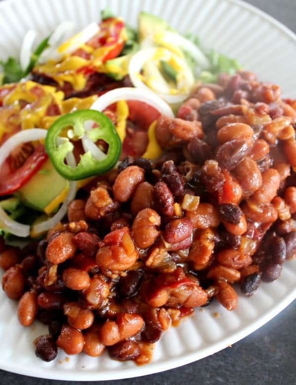Best Baked Beans on white plate with burger fixings on side, tomatoes, mustard, onions, jalapeno