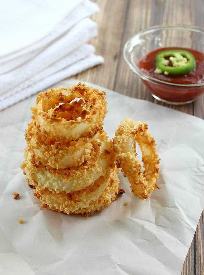 baked onion rings on parchment next to a bowl of ketchup with jalapeno