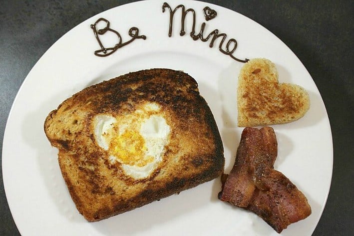Egg in the hole with Be Mine written in chocolate on plate and heart-shaped bacon and toast 