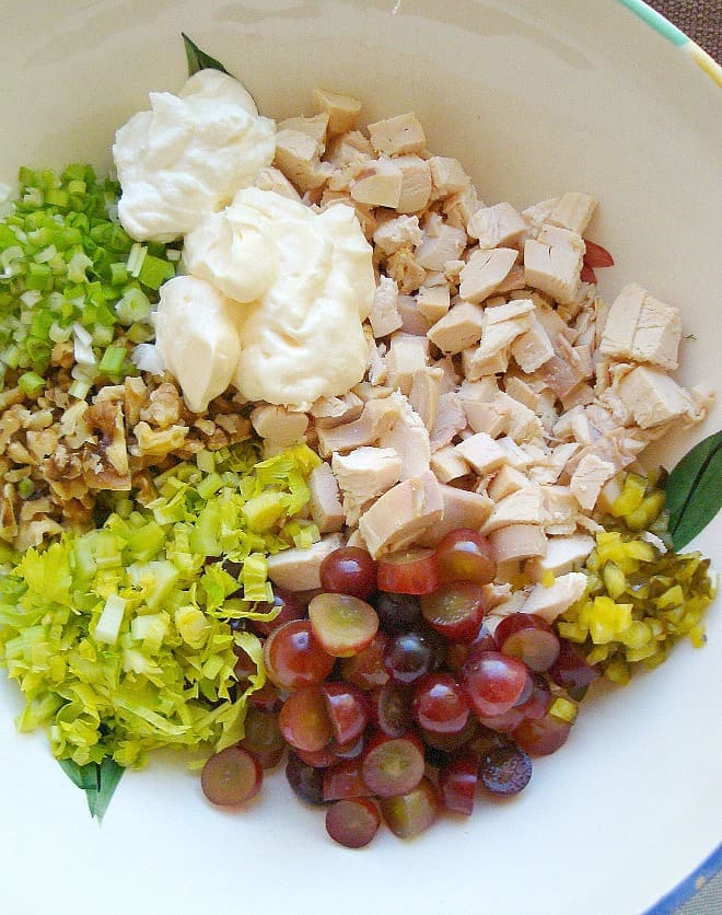 ingredients for chicken salad in a white bowl. Mayo, chicken, relish, grapes, celery, walnuts, scallions.