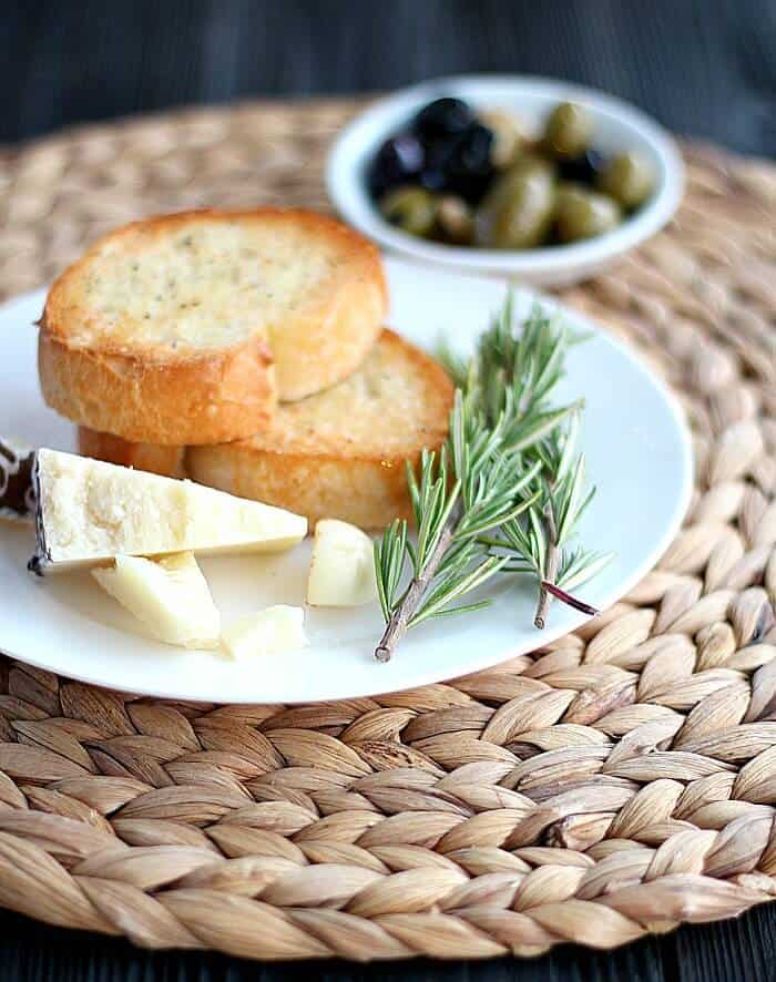 Toasted baguette, cheese, thyme on a white plate with a bowl of olives in the back