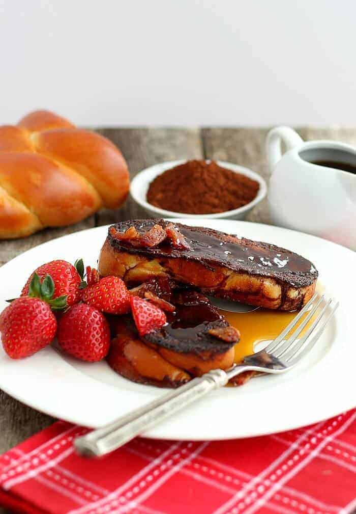 Chocolate French Toast on a white plate with strawberries. Chocolate and syrup in background and cut challah bread