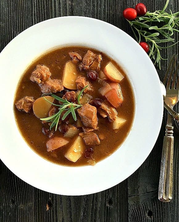 Cranberry Beef Stew in white bowl with rosemary on top, utensils to side