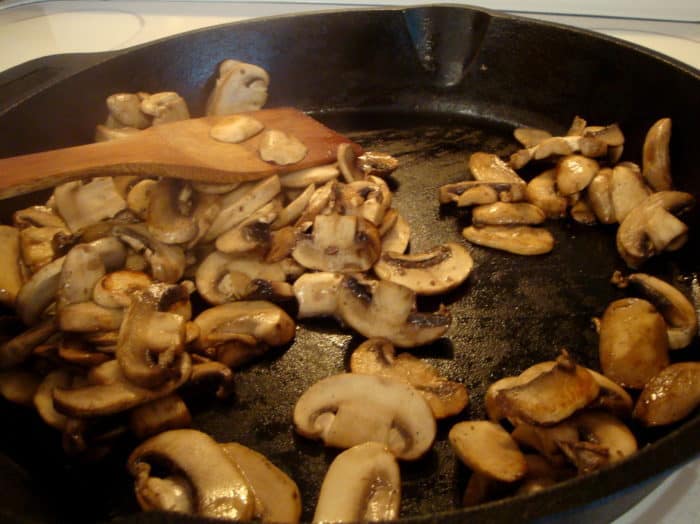 sliced mushrooms cooking in a black skillet, being stirred with a wooden spoon