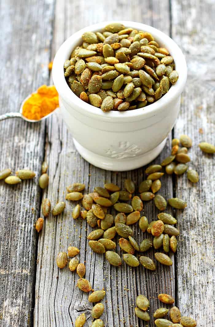 Turmeric Roasted Pumpkin Seeds in a white bowl with turmeric on a spoon and seeds on the side 